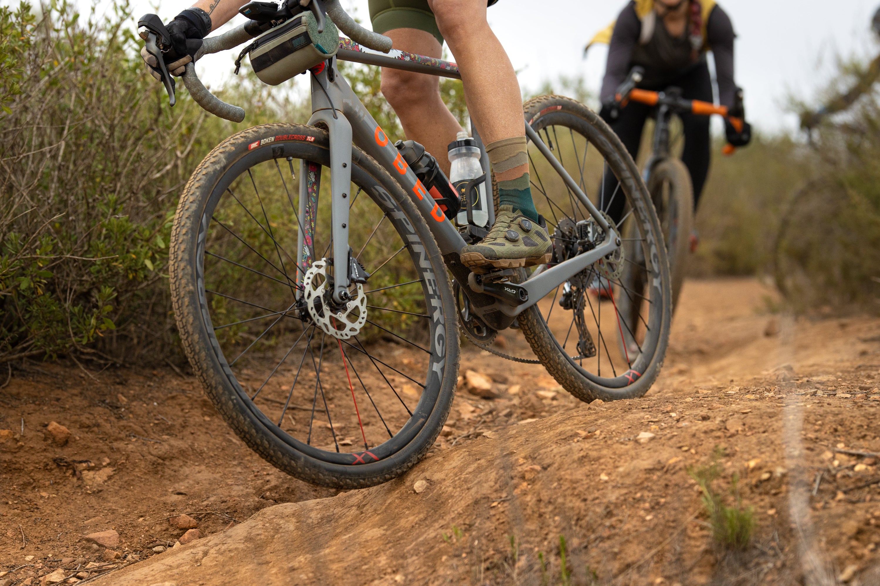 Two people OBED Gravel Performance bicycles on a dirt trail with a natural landscape.