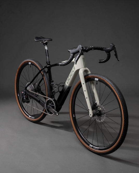 A high-end gravel bicycle with a matte black frame, a white front fork, and brown sidewall tires, photographed in a studio setting against a dark gray background.