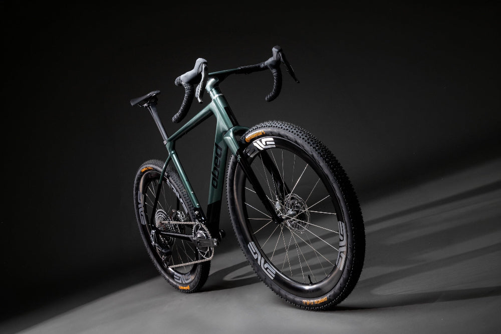 A gravel bicycle with a dark metallic green frame, black components, and knobby tires, photographed in a dramatic studio setting with a dark gray background.