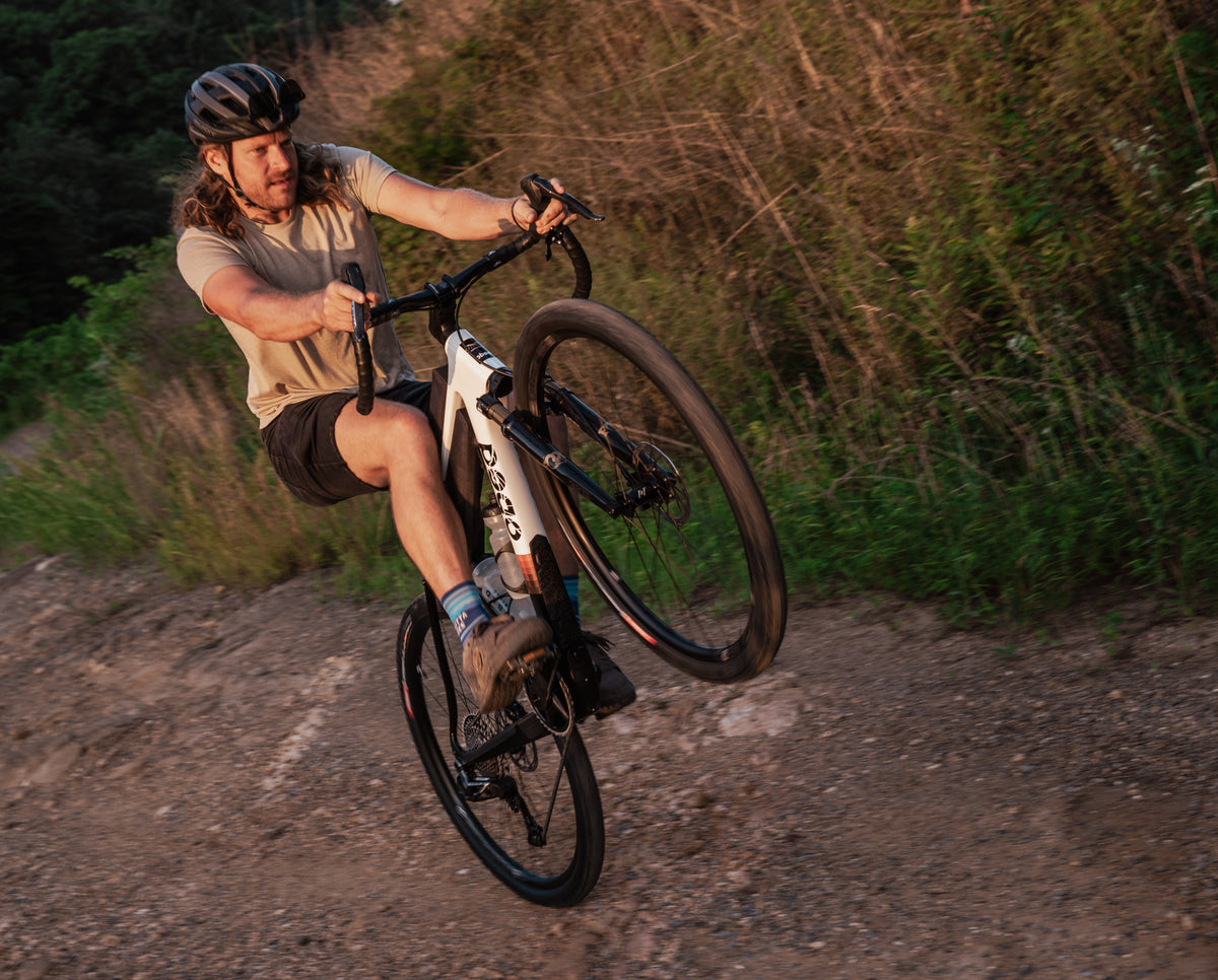 A man wearing a helmet and tan t-shirt rides a Parallax MMR bike on a dirt trail outdoors during golden hour, with green brush in the background.