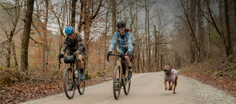 Two people riding OBED Boundary bikes on a gravel road with dog running beside	