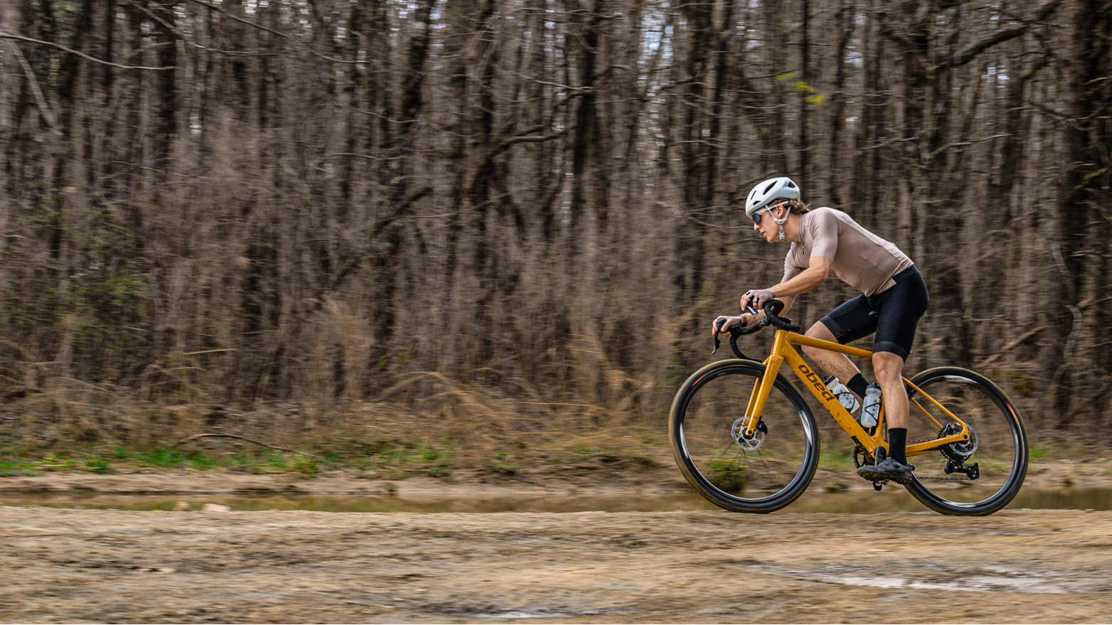 Person riding a yellow bicycle on a dirt path with leafless trees in the background